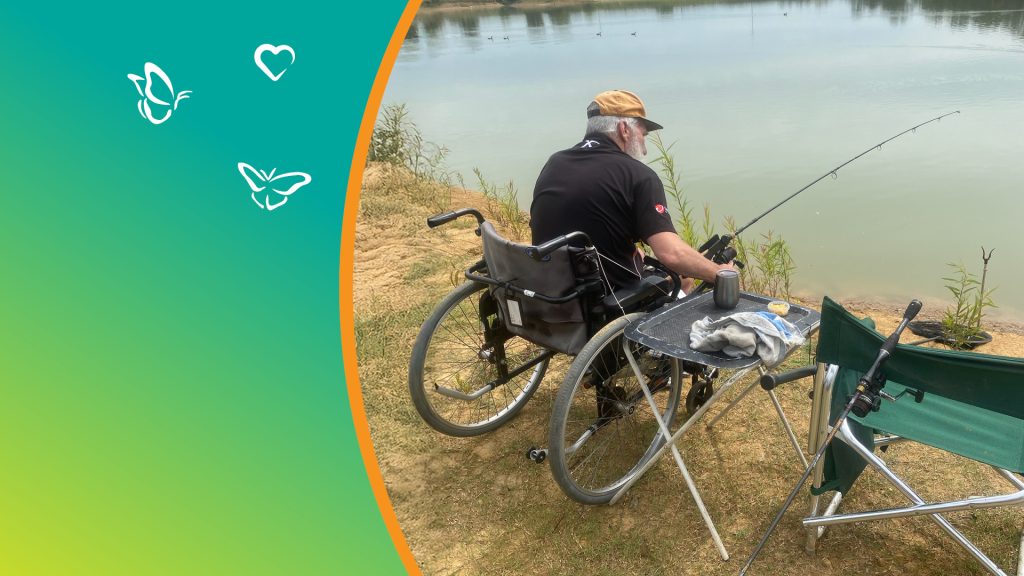An older man sits in a wheelchair at the edge of a calm lake, fishing quietly. He wears a tan cap and a black shirt, holding a fishing rod extended over the water. A small folding table beside him holds a cup, a cloth, and a snack, with another fishing rod resting near a green chair. The scene is peaceful and inclusive, framed by a teal and green gradient design with white butterfly and heart icons along the left side, symbolising serenity and care.