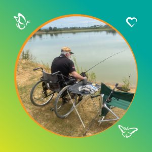 An older man sits in a wheelchair at the edge of a calm lake, fishing on a quiet day. He wears a tan cap and a black shirt, holding a fishing rod extended over the water. A small folding table beside him holds a cup, a cloth, and a snack, with another fishing rod resting near a green chair. The scene is framed by a circular border with a green-to-yellow gradient and small butterfly and heart icons, evoking a peaceful, inclusive moment outdoors.