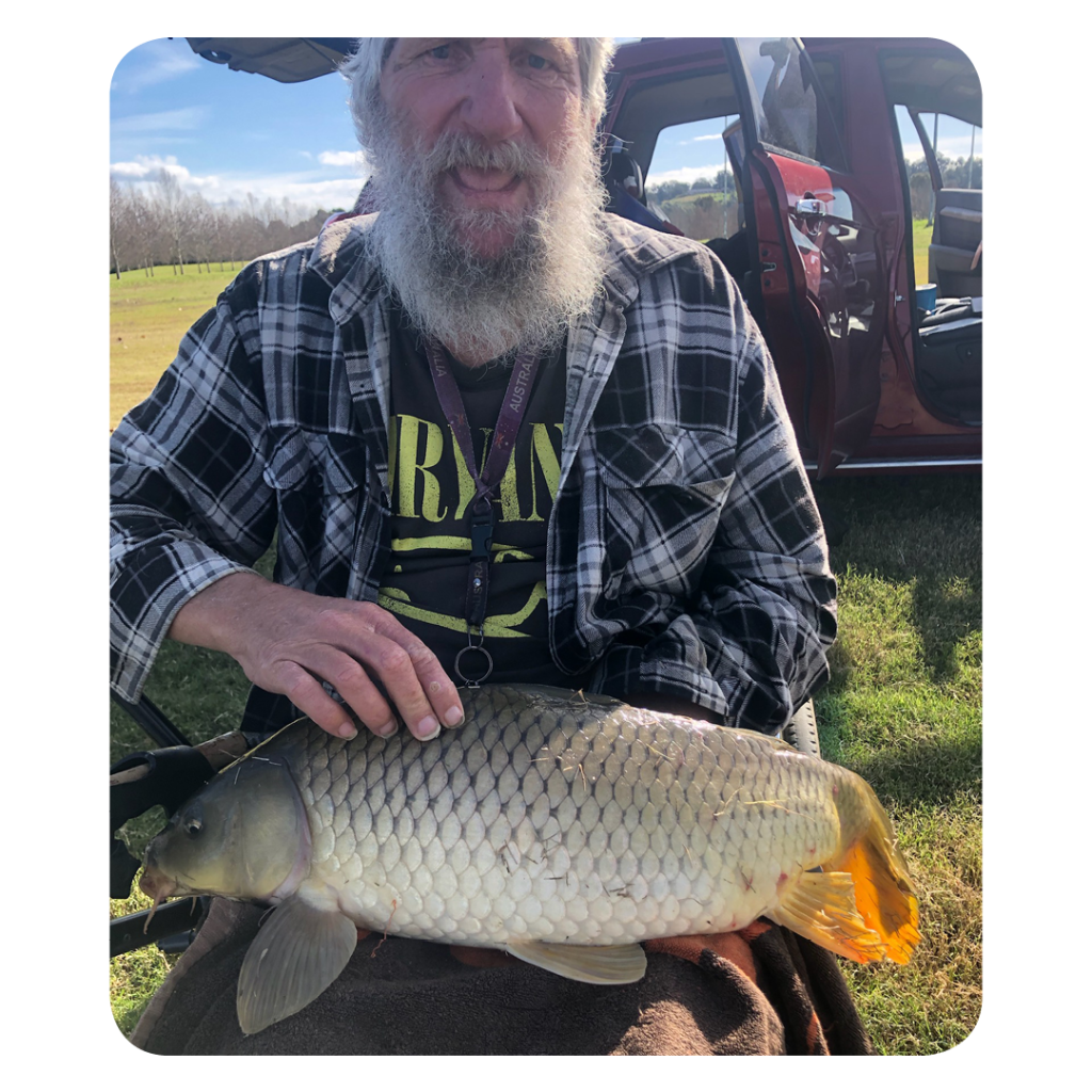An older man with a long gray beard smiles while holding a large carp across his lap. He wears a black-and-white plaid shirt over a Nirvana T-shirt and has a lanyard around his neck. Behind him, a red vehicle with its door open sits on a grassy field under a bright blue sky with scattered clouds. The scene conveys pride and contentment after a successful day of fishing.