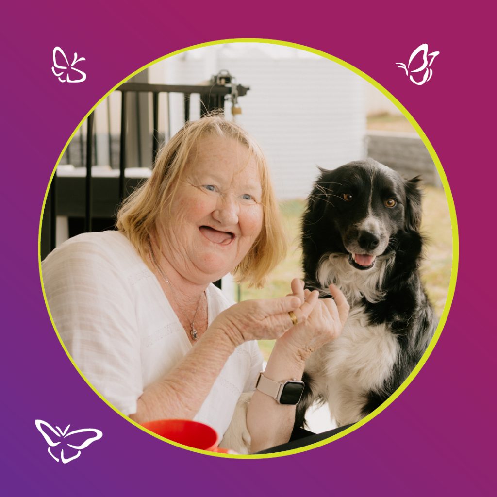 An older woman with light hair and a joyful smile sits beside a black and white dog. She wears a white blouse and a smartwatch, holding her hands near the dog as they both look toward the camera. The photo is framed in a circular border with a yellow outline against a purple-to-magenta gradient background decorated with white butterfly illustrations, creating a warm and cheerful atmosphere.