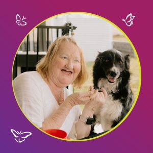 An older woman with light hair and a joyful smile sits beside a black and white dog. She wears a white blouse and a smartwatch, holding her hands near the dog as they both look toward the camera. The photo is framed in a circular border with a yellow outline against a purple-to-magenta gradient background decorated with white butterfly illustrations, creating a warm and cheerful atmosphere.