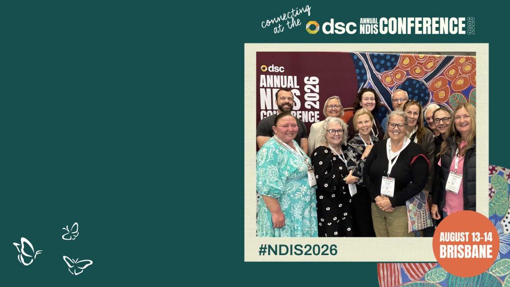 A group of twelve smiling conference attendees stand together in front of a colorful abstract backdrop featuring circular and organic patterns in red, blue, orange, and green. They wear name badges and lanyards, posing for a photo at the DSC Annual NDIS Conference 2026. The image frame includes text reading “connecting at the DSC Annual NDIS Conference 2026,” “#NDIS2026,” and “August 13–14 Brisbane,” with small butterfly icons on a dark green background.