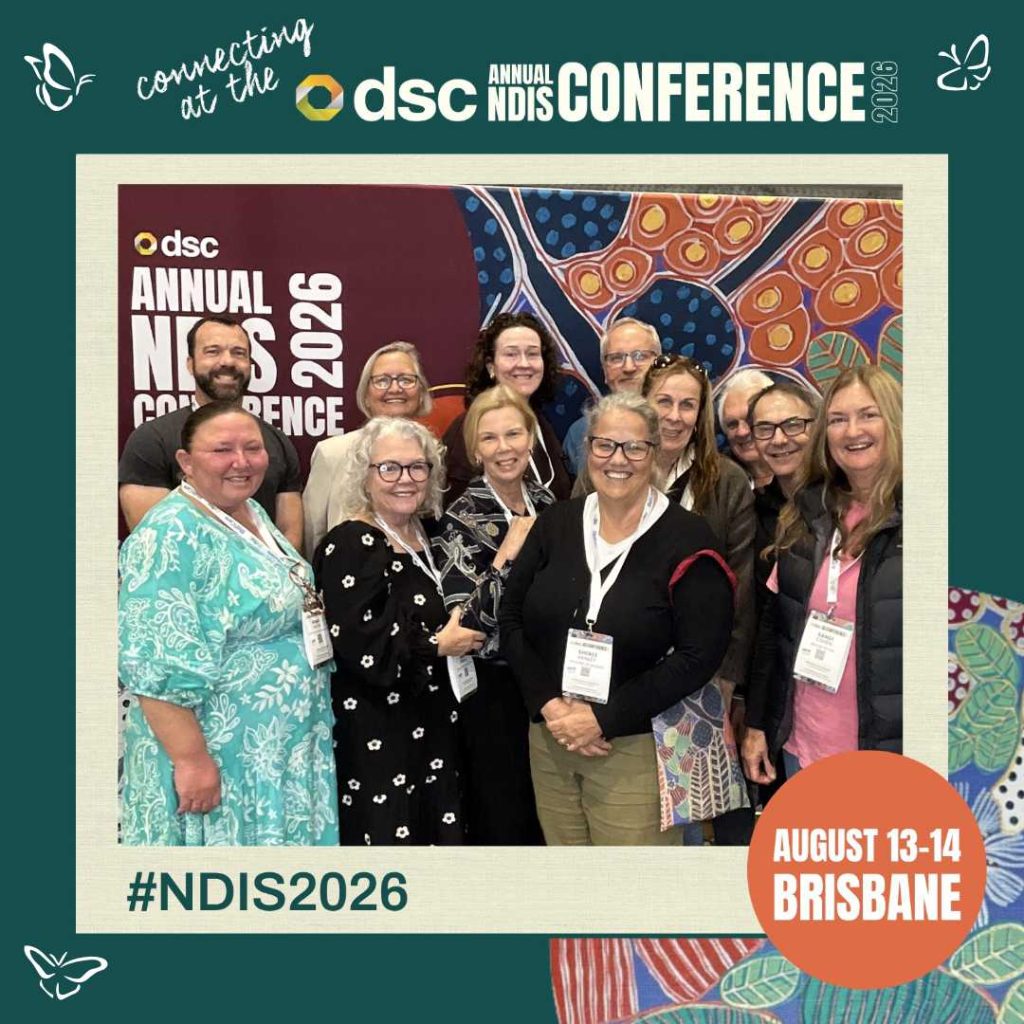 A group of twelve smiling conference attendees stand together in front of a colorful abstract backdrop featuring circular and organic patterns in red, blue, orange, and green. They wear name badges and pose for a photo at the DSC Annual NDIS Conference 2026. The frame around the photo includes text reading “connecting at the DSC Annual NDIS Conference 2026,” “#NDIS2026,” and “August 13–14 Brisbane,” with small butterfly icons in the corners.