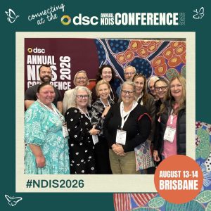 A group of twelve smiling conference attendees stand together in front of a colorful abstract backdrop featuring circular and organic patterns in red, blue, orange, and green. They wear name badges and pose for a photo at the DSC Annual NDIS Conference 2026. The frame around the photo includes text reading “connecting at the DSC Annual NDIS Conference 2026,” “#NDIS2026,” and “August 13–14 Brisbane,” with small butterfly icons in the corners.